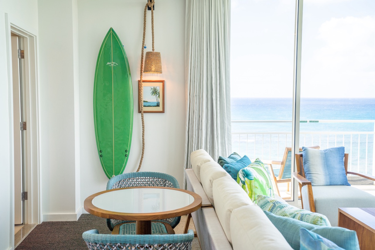 Green surf board leaned against wall, beside a hanging lamp and picture with dining table in front