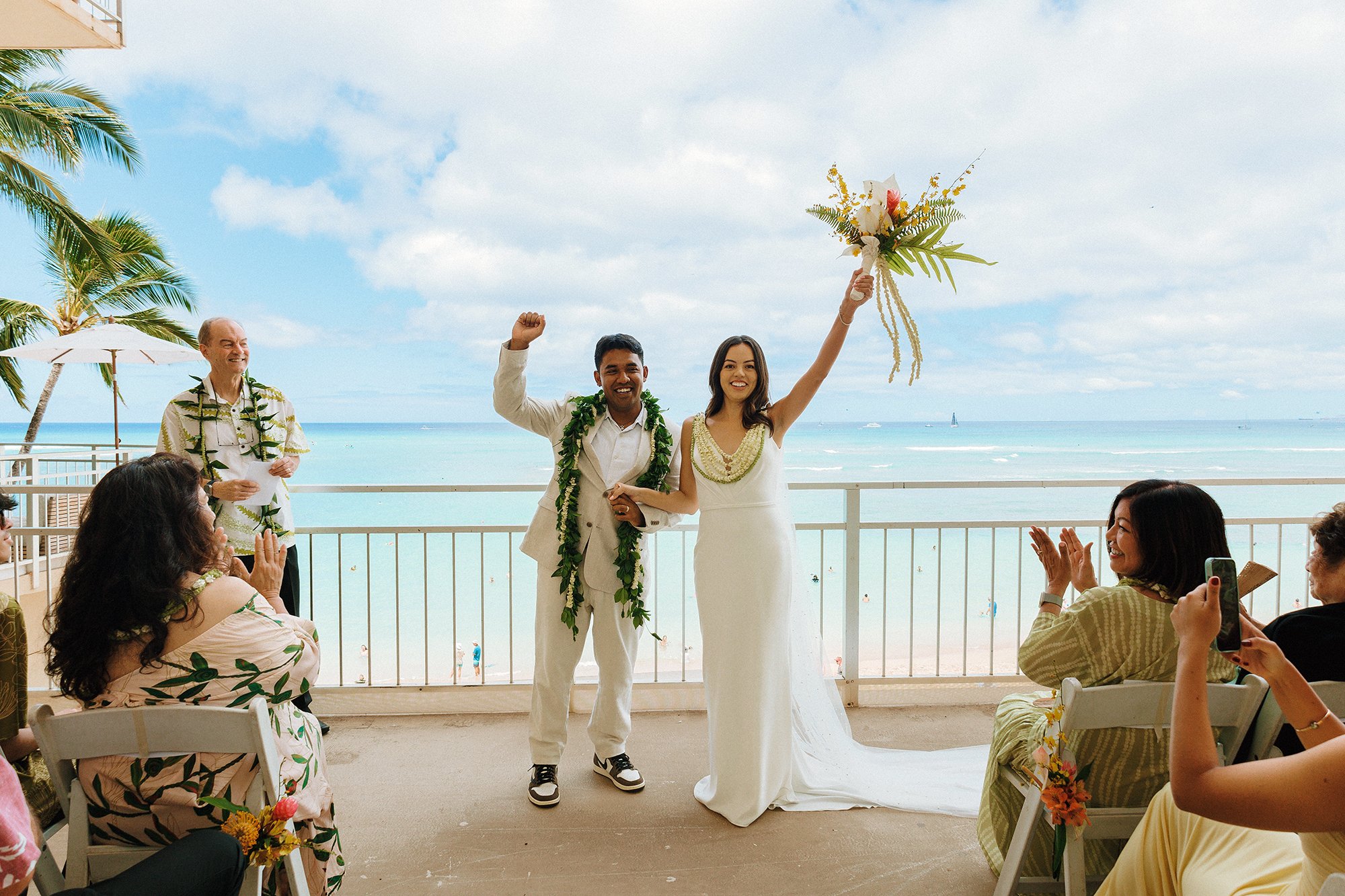 The happy bride and groom pump their hands in the air in celebration, the bride holds a bouquet of flowers